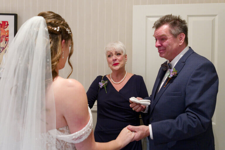 The brides parent see her in her wedding gown for the first time. They look happy and emotional.