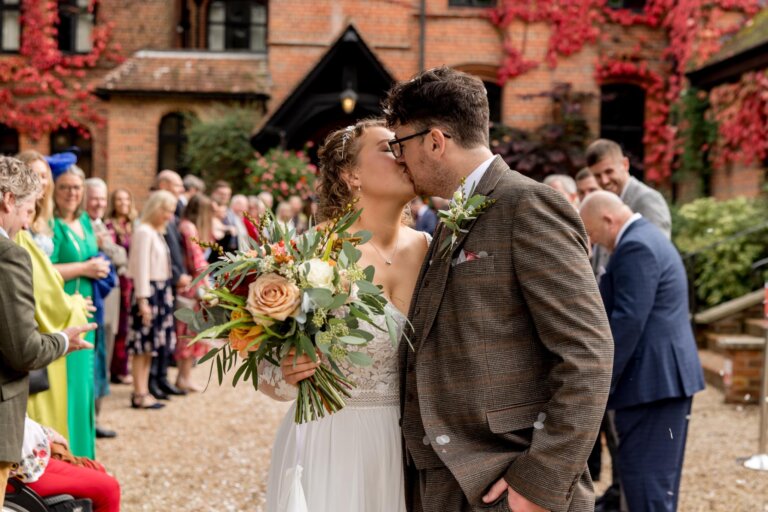 Bride and Groom share a kiss with guests and Careys Manor in the background